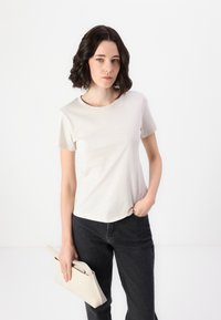 Short-sleeve, cream-colored t-shirt with a round neckline, paired with dark jeans. The model holds a light beige clutch. Simple and casual design.