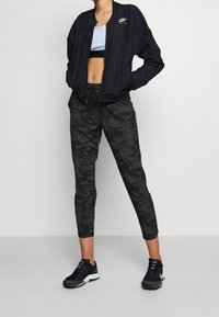 Svart bomberjacka med Nike-logga, en kort blå topp och mörka camo-joggingbyxor. Svarta sneakers fulländar den sportiga looken.