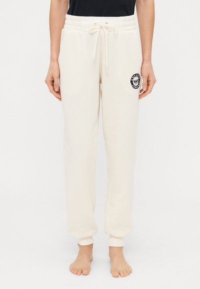 Cremefarvede sweatpants lavet af blødt stof, med elastisk talje, snøre og et cirkulært logo på venstre lår.