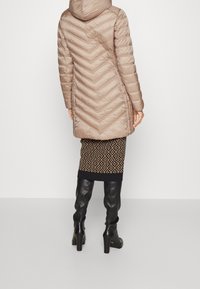 Beige-värinen tikkitakki hupulla, jossa on kalanruotokuvio. Päällä musta hame, jossa on geometrinen kuvio, ja polvenpituiset mustat saappaat.