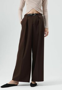 Wijde bruine broek met plooien aan de voorkant, gemaakt van soepel stof. Gecombineerd met een nauwsluitend licht topje en zwarte instapschoenen.