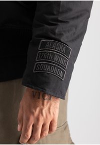 Zwarte jacksleeve met drie geborduurde patches: "ALASKA," "176e VLEUGEL," en "SQUADRON." Het materiaal lijkt glad met een ribgebreide manchet.