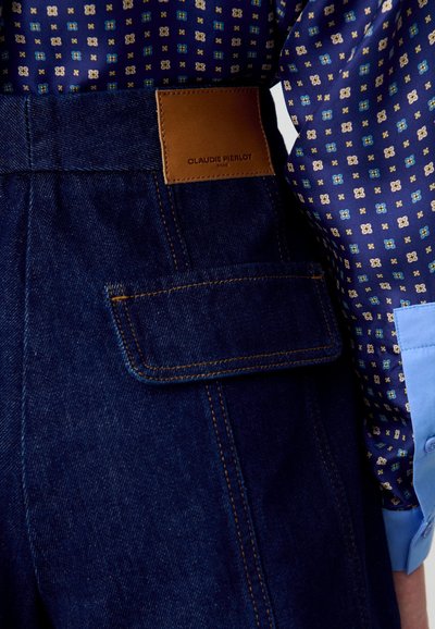 Pantalones de mezclilla oscuros de tiro alto con una etiqueta de cuero marrón que dice "Olaudie Pierlot", que presentan un bolsillo con solapa y costuras en contraste.