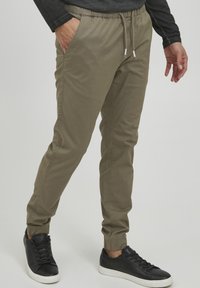 Olijfgroene joggers met een elastische tailleband en trekkoord, met zijzakken en elastische boorden. Gecombineerd met zwarte sneakers.