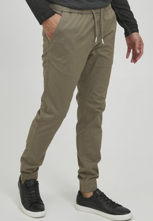 Olivgrüne Jogginghosen mit elastischem Bund und Kordelzug, seitlichen Taschen und elastischen Bündchen. Getragen mit schwarzen Turnschuhen.