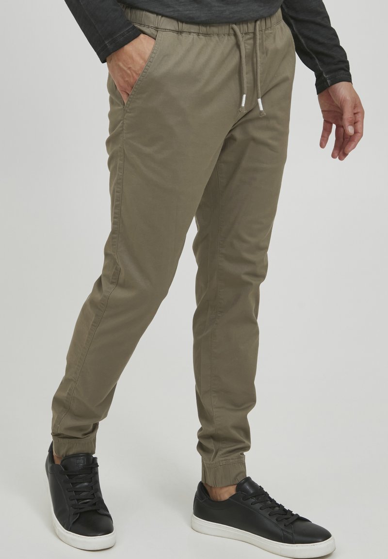 Olijfgroene joggers met een elastische tailleband en trekkoord, met zijzakken en elastische boorden. Gecombineerd met zwarte sneakers.