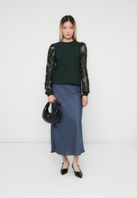 Felpa verde scuro con maniche in pizzo floreale trasparente, abbinata a una gonna midi in satin blu e una borsa nera, con scarpe nere a punta.