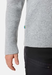 Close-up of a person’s left hand wearing a light gray ribbed sweater and black pants with a zipper pocket on the side.