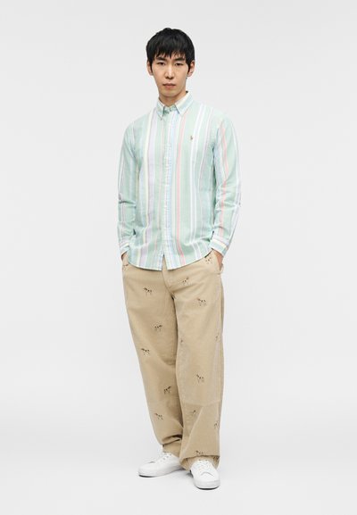 Ανοιχτό πράσινο ριγέ πουκάμισο με κουμπιά, συνδυασμένο με ταμπά φαρδιά παντελόνια που έχουν κεντημένα μοτίβα σκύλων. Λευκά αθλητικά παπούτσια ολοκληρώνουν την εμφάνιση.