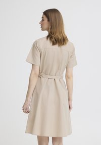 Vrouw met schouderlang lichtbruin haar, gekleed in een beige jurk met korte mouwen en een ceintuur, gezien vanaf de achterkant tegen een effen achtergrond.