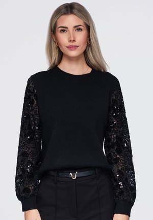 Une femme porte un pull noir aux manches transparentes brodées de sequins, assorti à un pantalon noir et à une ceinture, sur un fond uni.