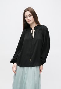 Čierna blúzka s dlhým rukávom, výstrihom v tvare kľúčovej dierky, zdobená nariasením a zlatým príveskom hviezdy na šnúrke. Mäkká, hladká tkanina.