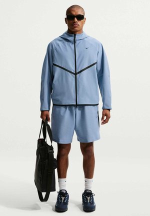 Hombre con chaqueta deportiva azul claro y pantalones cortos, calcetines blancos, zapatillas oscuras, gafas de sol, sosteniendo una bolsa negra sobre un fondo liso.