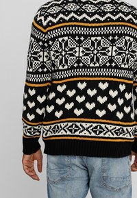 Maglione nero lavorato a maglia con motivi bianchi e gialli, caratterizzato da motivi a forma di cuore e design geometrici. Tessuto spesso, polsini e orlo a coste.