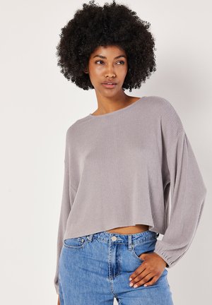 Pull long à manches longues gris clair, côtelé, avec un large col et un ourlet cropped, associé à un jean bleu taille haute. Texture douce, coupe décontractée.
