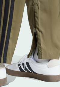 Pantalones de chándal verde oliva con rayas marinas, que cuentan con aberturas laterales con cremallera, combinados con zapatillas blancas y acentos gris claro.