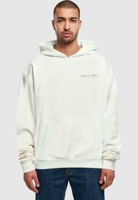 Weißer Oversize-Hoodie mit einer Vorderseite Tasche, aus weichem Material und besticktem Text in Grün. Kombiniert mit dunkelblauen Jeans.