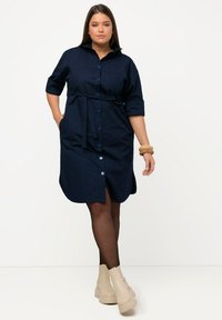 Abito button-up blu navy con colletto, maniche corte, vita con cintura e spacco sul fondo. Indossato con stivaletti beige.