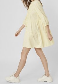 Gul, lager-på-lager klänning i lätt tyg med långa ärmar och dragsko vid halsringen; ihop med vita sneakers.