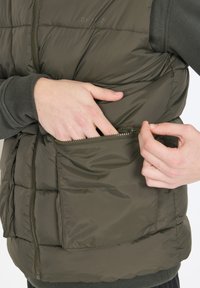 Dunkelgrüne Steppweste mit gep quilteter Textur, ausgestattet mit einer Frontreißverschlusstasche und einem seitlichen Reißverschluss. Das Material wirkt leicht und isoliert.