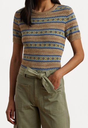 Vrouw draagt een gebreide trui met korte mouwen met bruine, blauwe en groene strepen, gecombineerd met olijfgroene broek met hoge taille en een gestrikte riem.