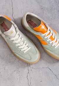 Paire de baskets décontractées en daim vert clair, lacets blancs, semelles en gomme et accents en cuir orange contrastants sur une chaussure, sur un sol en béton fissuré.