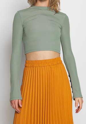 Frau trägt ein salbeigrünes, langärmeliges, kurzes Oberteil zusammen mit einem hochtaillierten, plissierten, leuchtend orangen Rock vor einem schlichten Hintergrund.