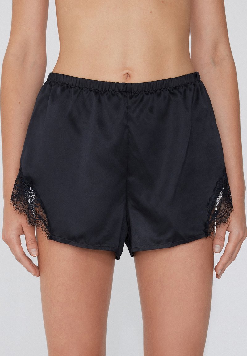 Zwarte satijnen shorts met een elastische tailleband, voorzien van kantaccenten aan de zoom. Soepele textuur, relaxte pasvorm en knielange ontwerp.