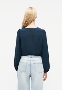 Ξανθός/ή με την πλάτη, φορά μπλούζα μακρυμάνικη σκούρου μπλε χρώματος με φουσκωτούς ώμους και παντελόνι τζιν ανοιχτού μπλε, ψηλόμεσο.