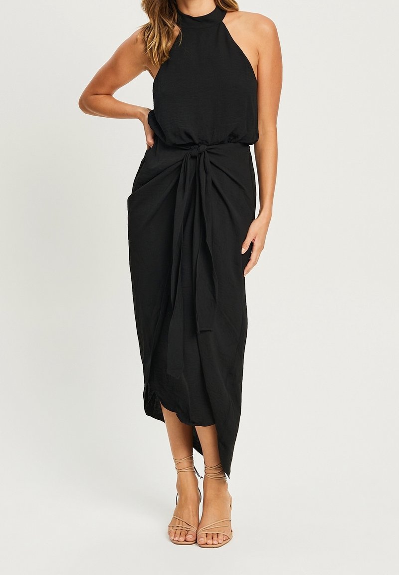 Robe noire à col halter en tissu léger. Caractérisée par un détail de taille nouée et une longueur midi. Le modèle porte des sandales beiges à lanières.
