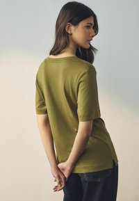 Olijfgroen kortmouwshirt met een ronde hals, zachte textuur, zijsplitten en een ontspannen pasvorm, gecombineerd met donkere jeans.