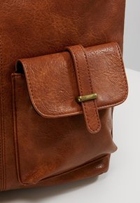 Brun lædertaske med lomme og klap sikret af en rem og messing spænde, der viser detaljeret tekstur og syning på en neutral baggrund.