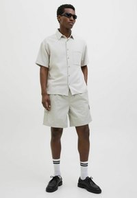 Chemise gris clair à manches courtes avec une poche, associée à un short cargo gris clair. Des lunettes de soleil noires et des chaussures noires avec des chaussettes rayées blanches complètent le look.