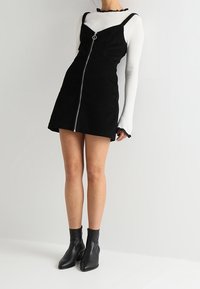 Svart manchestertklänning med dragkedja fram och metallringdetalj, bärs över en vit ribbad långärmad topp. Svarta ankelboots fulländar looken.