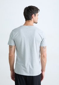 T-shirt di cotone grigio, maniche corte, scollo rotondo, texture liscia, design aderente, indossato con pantaloni neri, tatuaggi visibili sulle braccia.
