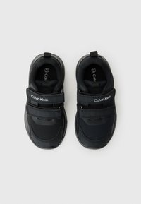 Chaussures de sport noires avec une tige en textile lisse, des bandoulières Velcro et des semelles en caoutchouc avec des motifs texturés. Comprend le logo de la marque sur les bandoulières.