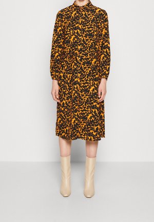 Kvinde iført en langærmet, orange og sort blomstrede midikjole med krave, kombineret med cremefarvede ankelstøvler.