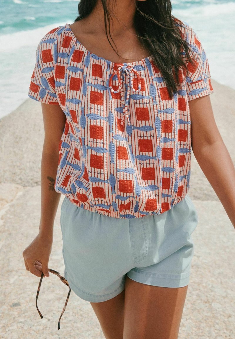Femme portant un chemisier à manches courtes rouge et bleu à motifs et un short bleu clair, tenant des lunettes de soleil près d'une jetée de plage.