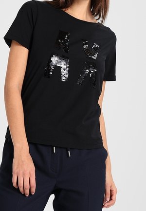 Kobieta ubrana w czarną koszulkę z krótkim rękawem, z przodu znajduje się abstrakcyjny wzór z połyskujących czarnych i srebrnych cekinów.