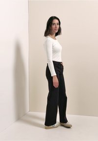 Haut blanc à manches longues associé à un pantalon noir taille haute à jambes larges, doté d'un design simple et d'une ceinture orange-brun, sur un fond neutre.