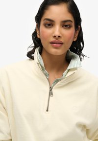 Sudadera color crema con cuello con cremallera y forro interior de rayas azul claro, que presenta un logo sutil en el pecho.