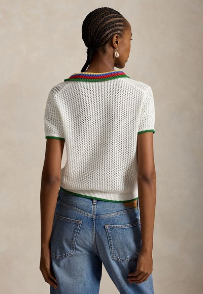 Γυναίκα με πλεγμένα μαλλιά που φοράει λευκή πλεκτή κοντομάνικη μπλούζα με πολύχρωμο γιακά και μπλε τζιν, στέκεται με την πλάτη στραμμένη σε απλό φόντο.