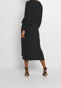 Schwarzes Langarmkleid mit Peplum-Taillierung, mittellangem Saum und glatter Textur, ergänzt durch hohe Schuhe mit Knöchelriemen.