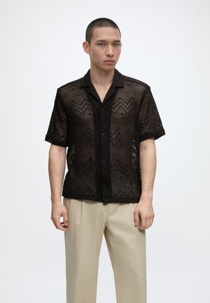 Młody mężczyzna ubrany w czarną siateczkową koszulę z krótkim rękawem z wzorem zygzakowym i beżowe spodnie, stojący na jednolitym białym tle.