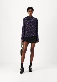 Black shirt with purple floral pattern, long sleeves, and a high collar. Paired with black shorts and chunky heeled sandals. Olive green bag.