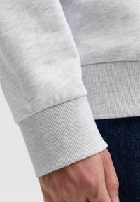 Tejido de sudadera de color gris claro con una textura suave y puños acanalados. Primer plano de una mano apoyada sobre un fondo de tela de mezclilla.