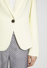 Jasnożółta marynarka z jednym brązowym guzikiem, o dopasowanym kroju i gładkiej fakturze, zestawiona z spodniami w srebrny wzór o fakturze.