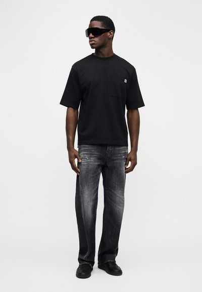 Μαύρο T-shirt με κοντό μανίκι και τσέπη στο στήθος, συνδυασμένο με ξεβαμμένο γκρι τζιν με φθαρμένες λεπτομέρειες και σκούρα παπούτσια.