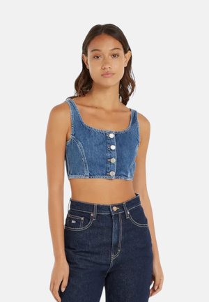 Giovane donna che indossa un crop top di denim blu senza maniche con bottoni frontali e jeans blu scuro a vita alta su uno sfondo semplice.