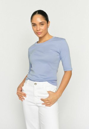 Woman with slicked-back hair wearing a light blue elbow-length sleeve top and white high-waisted pants, posing with hands in pockets against white background.